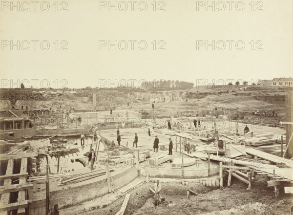 Construction of Locks at Veere. Possibly No. 10 Lock chambers and inner heads of the double Lock at Veere, viewed from the pier between the outer heads, Veere, lock under construction, photograph, photographer: Pieter Oosterhuis, 1871-08, pasteboard, paper, albumen print, height 320 mm x width 419 mm, weigh-house, A wide construction site with exposed foundations, wooden planks, beams, and scaffolding stretching across the foreground and middle distance, with workers scattered on platforms and among the construction materials; shallow water pooled in trenches and channels near the front, piles of stone and rubble, and various tools and carts visible; earthworks and mounds of soil rise toward the background where stacks of logs and stone blocks sit behind a low ridge; a small grove of trees and a few houses with pitched roofs appear on the horizon; the sky above is pale and mostly empty.