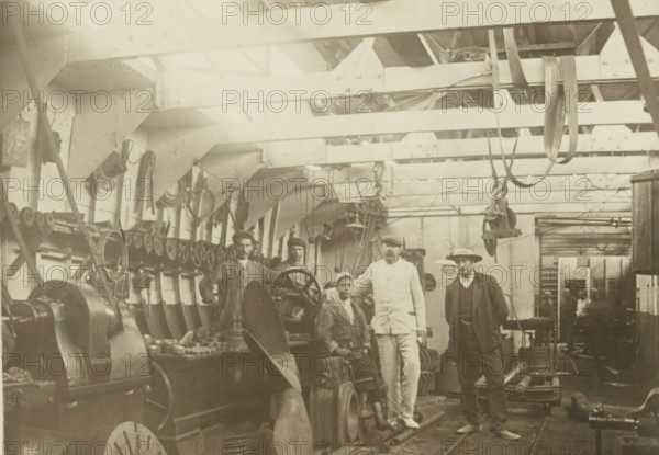 Machine shop, photograph, Interior of a machine shop. Photo pasted on cardboard, without texts. Part of the folder with 44 photos of dredgers from the company A. Bos & JP Heijblom in Dordrecht., c. 1900 - c. 1910, photographer, paper, pasteboard, gelatin silver print, developing gelatin silver print pasted on cardboard, height 122 mm x width 171 mm, height 240 mm x width 298 mm, Person, Firma A. Bos & JP Heijblom, factory interior, dredge, dredging machine, A sepia-toned interior shows a workshop with overhead beams and hanging straps and pulleys, a row of mounted wheels and machinery along the left wall, large gears and a propeller blade in the foreground, workbenches with tools and parts, and five men standing and seated near the machines wearing work clothes, a suit with coat and hat, and a light-colored uniform; the ceiling has structural supports and lighting, and the background contains more equipment and a doorway leading to another room.