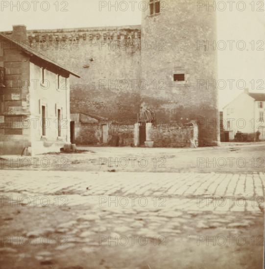 View in an unknown city, France, photograph, France., c. 1900 - c. 1903, photographer, paper, daylight gelatin silver print, height 84 mm x width 82 mm, public road in village, social history, population, Period, 1875 - 1925, A wide cobbled street fills the foreground leading to an open area bordered by a tall textured stone wall and a round stone tower with a small barred window, adjacent to a lower building with light-colored façades and dark roof eaves; remnants of a low stone barrier and an open gateway sit at the base of the tower, and a smaller multi-story building with pale walls and multiple windows is visible further to the right.