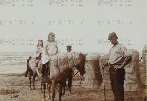 At the Beach, photograph, inscription: 'H + Nan, zeev, ...', photographer: attributed to Willem de Jong, Netherlands, photographer: attributed to Piet de Jong, c. 1900, Attributed to, paper, baryta paper, daylight collodion silver print, height 90 mm x width 120 mm, Person, Johanna Elisabeth Borgerhoff Mulder-Vreede (Nan), Johanna Petronella Lambertina Stokhuyzen-de Jong (Hans), beach, riding a horse, ass, or mule, rider, horseman, child, North Sea, A sepia-toned beach scene shows two young girls sitting on horses facing forward, each wearing light dresses and patterned headscarves, with one girl nearer the camera looking down and the other slightly behind smiling. A man stands to the right wearing a cap, a long-sleeved shirt with sleeves rolled, striped trousers, and holding a walking stick while looking toward the camera. Another man in a dark cap stands between the horses, partially obscured. The horses have light manes and saddles, and large woven wicker baskets or beach chairs sta...