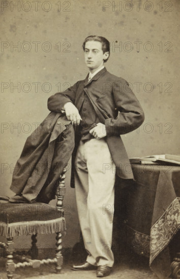 Portrait of an Unknown Young Man, carte-de-visite, name: 'Wegner & Mottu photo. Amsterdam', photographer: Wegner & Mottu, Amsterdam, c. 1865, height 89 mm x width 57 mm, height 105 mm x width 62 mm, historical persons not known by name, A sepia-toned portrait of a young man standing with one elbow resting on the back of an upholstered chair draped with a dark coat, wearing a dark jacket over a lighter waistcoat and light-colored trousers, his hair neatly combed back and his expression neutral, one hand resting on his hip and the other holding the coat, a small table covered with a dark cloth and a folded cloth with a patterned border visible beside him, and the background showing a plain mottled backdrop.
