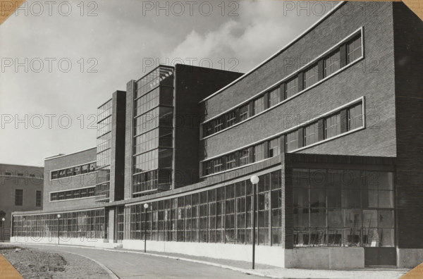 Italy, photograph, c. 1880 - c. 1910, photographer, paper, albumen print, height 300 mm x width 200 mm, A black and white photograph of a multi-story building with a curved facade and long horizontal bands of windows set into brickwork; large vertical glass stairwell sections project from the front and reveal a grid of window panes and interior stair treads; the ground level features a continuous row of tall glass panels framed by slender mullions, some showing reflections and interior shelving; simple street lamps with spherical globes line the sidewalk in front of the building; the sky above is cloudy and light in tone; the foreground shows a paved road curving past a grassy verge.
