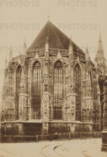 St. Sebald Nürnberg, photograph, c. 1850 - c. 1900, photographer, paper, albumen print, albumendruk, height 162 mm x width 104 mm, height 182 mm x width 134 mm, Bavaria, Nuremberg, St. Sebaldus Church, A sepia-toned photograph of a large Gothic church exterior showing a pointed tiled roof and tall ornate windows with pointed arches, tracery, and mullions; the stone facade features vertical buttresses, pinnacles, carved niches with statues, decorative stone carvings, and sculpted details; a rounded spire is visible to one side, and temporary wooden fencing runs along the base of the building in front of a stone plinth; the foreground shows a curved roadway with faint tram tracks and a cylindrical street fixture at the near right edge.