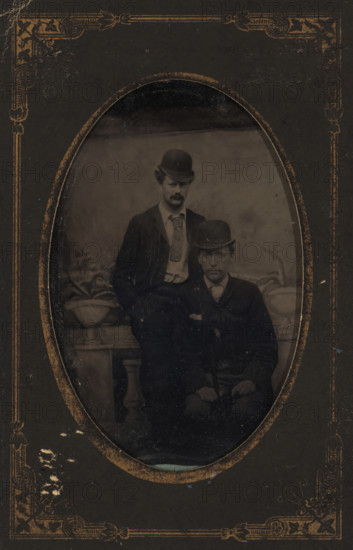 Portrait of Willem Kloos and Hein Boeken, photograph, photographer: unknown, Netherlands, c. 1885 - c. 1890, metal, inorganic material, iron (metal), paper, ferrotyping, ferrotype in paper cover, height 81 mm x width 53 mm, height 118 mm x width 78 mm, Person, Willem Kloos, Hein Boeken, historical persons, head-gear: hat, An oval-framed black-and-white photograph set within a dark decorative mat shows two men wearing dark suits and bowler hats; one man stands behind the other who is seated, both with neutral expressions, the standing man has a patterned necktie and light-colored shirt visible beneath his jacket while the seated man wears a darker necktie and holds a cane across his lap, a potted plant on a pedestal appears beside them and a plain backdrop fills the space behind, the mat around the oval has worn gold ornamental corner designs and scuffed edges.