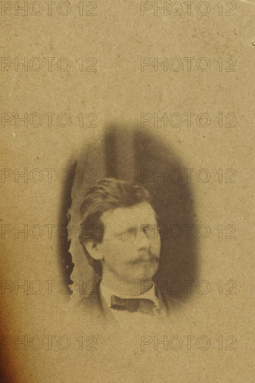 Portrait of Theodorus Kroes, photograph, number: '95', photographer: attributed to Antoon Bauduin, 1862 - 1866, Attributed to, paper, albumen print, albumendruk, height 99 mm x width 72 mm, historical persons (portraits and scenes from the life), A faded sepia-toned photograph showing the upper portion of a man's head and shoulders emerging from a darker oval vignette against a lighter background, with the man wearing a dark bow tie and a high-collared shirt, short wavy hair, and a mustache; the left edge of the image has a torn, darkened area with irregular burn-like edges and a rough white torn corner.