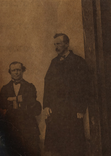 Self-portrait with unknown man (left), Two white men, one sitting one standing (former title), photograph, number: '73', photographer: attributed to Antoon Bauduin, 1862 - 1866, Attributed to, paper, albumen print, height 135 mm x width 92 mm, historical persons not known by name, A sepia-toned photograph shows two men standing against a plain backdrop, one shorter man seated or positioned slightly lower on the left wearing a dark suit with a bow tie and holding an object at chest height, and a taller man on the right wearing a dark coat with hands resting near his sides; the image has dark irregular burn-like edges and visible creases and discoloration across the surface.