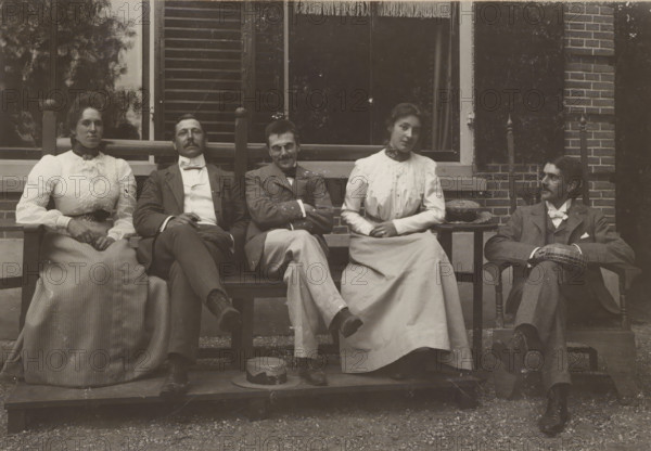 Group portrait with Hedwig Kleintjes, Gerard Aalbersberg, Jan Kleintjes and an unknown man and woman, photograph, Heerde (possibly), c. 1900 - c. 1906, paper, gelatin silver print, developing gelatin silver print, height 0 mm x width 0 mm, Five people sit on wooden chairs in front of a building with a window and brickwork, all wearing formal clothing from an earlier era; two women wear long light-colored dresses with high collars and lace or ruffled details, and three men wear suits with bow ties or neckwear and neatly groomed facial hair. The central man and the man to his right sit with legs crossed and arms folded while the men at the ends sit more relaxed with hands resting on laps or on armrests; a straw hat rests on the wooden platform at their feet and a small round woven basket sits on a side table beside one woman. The background shows shuttered and curtained windows framed by wooden posts and decorative finials on the chair backs, and the ground in front of the platform is gr...