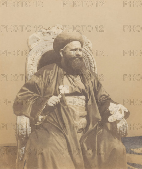 Portrait of a Coptic Patriarch in Cairo, photograph, inscription: '1859 La patriarche des Koptes au Kaire', inscription: '1082, 7 (M v W)', photographer: Willem de Famars Testas (possibly), Cairo (city), 1859, Ottoman Empire, North Africa, paper, paint (coating), deck paint, salted paper print, salt print, white body paint and drawn, height 186 mm x width 159 mm, historical persons not known by name, A seated man wearing a rounded head covering and a long dark robe is shown in a carved ornate chair; he has a full beard and a serious facial expression, one hand resting on the arm of the chair holding a small object and the other hand holding a folded patterned cloth, with a plain light background behind him.