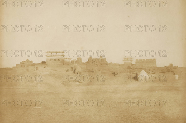 Luxor, View of Luxor, photograph, inscription: '1082, 7', inscription: 'Luxor', photographer: attributed to Ernest Benecke, 1852, North Africa, Attributed to, paper, salted paper print, height 140 mm x width 211 mm, city-view in general, 'veduta', A wide, faded sepia scene showing a low, uneven landscape with a distant line of ruined or weathered masonry structures and towers along the horizon, including a few square and cylindrical towers and sections of crenellated walls; in the foreground there is a flat, open expanse of ground with subtle undulations and faint marks, and toward the right foreground a small, light-colored domed hut with a low enclosing fence; the sky above is pale and empty.