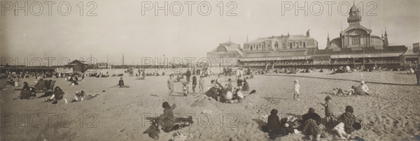 Beach on the North Sea coast, Belgium or France, panoramic photograph, Belgium, France, 1920 - 1940, paper, gelatin silver print, height 134 mm x width 385 mm, beach (as place of recreation), North Sea, A wide sandy beach scene with many people scattered across the sand, some sitting or reclining on blankets and towels, others standing or walking, and a few children playing near small mounds of sand; in the background a long seaside pavilion or complex of ornate buildings with large windows and a tower rises along the shoreline, with a covered veranda where more people are visible; beach equipment such as folding deck chairs and umbrellas appear among the visitors, and distant low structures and fencing line the horizon beneath a clear sky.
