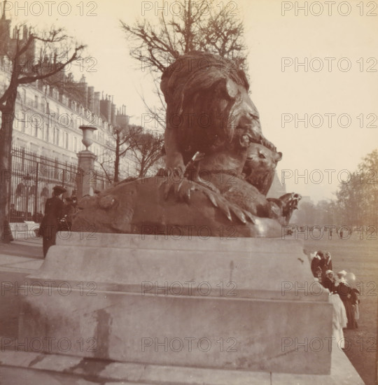 Tuileries, photograph, Paris, c. 1900, photographer, paper, pasteboard, baryta paper, daylight paper print, height 86 mm x width 82 mm, height 92 mm x width 90 mm, piece of sculpture, reproduction of a piece of sculpture, public gardens, park, A large bronze lion sculpture atop a stone pedestal fills the foreground, with the lion's head and mane visible in profile and one paw resting on another animal figure; behind the sculpture a row of leafless trees lines a paved promenade and an iron fence runs along the left side in front of tall multi-story buildings with chimneys; several people in period clothing and hats stand or walk near the fence and beside the pedestal, and a small group of figures is visible further down the promenade; the sky is light and mostly clear.