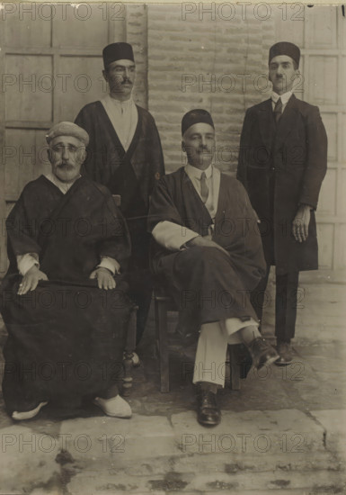 Portrait of Tavakoli (right) and 3 parliamentarians or ministers, photograph, Iran, 1900 - 1950, photographer, gelatin silver print, developing gelatin silver print, height 175 mm x width 127 mm, A sepia-toned portrait shows four men arranged in two rows against a brick and paneled background; two men sit in front on wooden chairs while two men stand behind them. The seated man on the left has a beard, wears a wrapped head covering and a dark robe with visible sleeve cuffs, and rests his hands on his lap where a dark fabric covers his knees; light-colored shoes peek out below his robe. The seated man on the right wears a dark brimless cap, a layered outfit with a lighter shirt and a tie, a draped outer garment, and has one leg crossed over the other with polished dark shoes. Behind them, the standing man on the left wears a brimless cap, a light shirt with a dark outer robe, and has a thin mustache. The standing man on the right wears a brimless cap and a dark suit with a high-collared...