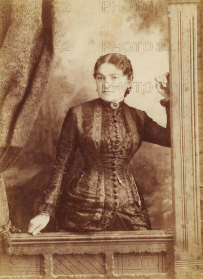 Portrait of Mrs JH McTavish from Winnnipeg, cabinet photograph, photographer: William Notman, photographer: Sandham, after 1867, paper, albumen print, albumendruk, height 165 mm x width 110 mm, A sepia-toned portrait of a woman shown from the hips up wearing a patterned, high-collared dress with a row of buttons down the front and a decorative brooch at the throat; her hair is styled and pulled back, and she has earrings. She is posed with one hand resting on a wooden ledge and the other hand gripping a vertical wooden frame, with patterned drapery visible on one side and a plain backdrop behind her. The image shows the texture of the fabric, the carved details of the wooden frame and ledge, and the overall worn, aged appearance of the photograph.