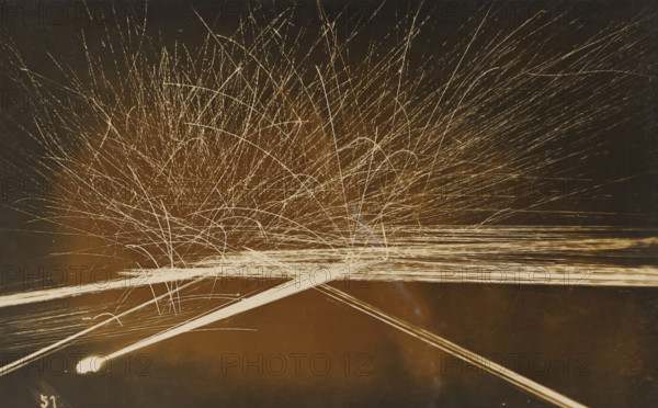 Barrage with machine gun(s) at evening or night, photograph, name: 'PHOTO WIDMER, WALLENSTADT', inscription: 'Tir de barrage, mitrailleuse, FM -, FJ', number: '51', photographer: E. Widmer, c. 1930, paper, gelatin silver print, developing gelatin silver print, height 88 mm x width 139 mm, A dark background filled with numerous thin, bright streaks radiating outward and upward from a lower central area, creating arcs and straight lines that intersect and overlap; a dense cluster of fine, light trails near the center spreads into longer, sparser streaks toward the edges; a few thicker, brighter linear bands run diagonally across the lower portion of the image, one ending in a small bright blob at the left side; overall tones range from warm brown to pale cream for the light trails against a deep, muted backdrop.