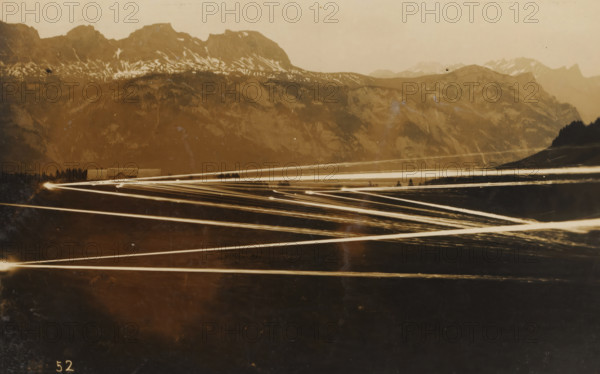 Barrage with machine gun(s) at evening or night, photograph, number: '52', name: 'PHOTO WIDMER, WALLENSTADT', inscription: 'Fauchage ?, horizontal…, mitrailleuse.', photographer: E. Widmer, c. 1930, paper, gelatin silver print, developing gelatin silver print, height 88 mm x width 139 mm, A sepia-toned landscape showing a range of mountains with patches of snow near their ridges, a band of forest at the foothills, and a low building with a peaked roof partially visible behind trees; across the foreground and middle ground multiple bright linear streaks of light cross the scene at various angles, converging toward bright points at the left and right edges, and the sky above the mountains appears hazy.