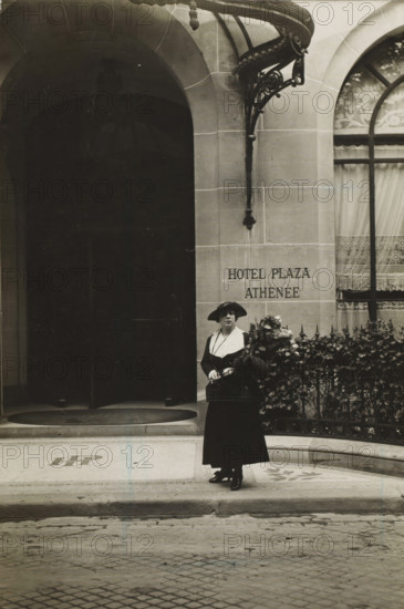 Woman on the street with a bouquet, photograph, inscription: 'Àffaire Costa', Paris, 1916, photographer, paper, gelatin silver print, developing gelatin silver print, height 117 mm x width 80 mm, A monochrome photograph of a woman standing on a tiled entrance in front of a stone building with arched openings; she wears a dark long skirt and jacket with a contrasting light collar and a wide-brimmed hat, holds a small handbag and a large bouquet of flowers, and stands beside an ornate iron railing covered with foliage; the building façade shows a decorative metal canopy and a curtained window with patterned fabric, and the left side of the image reveals a dark recessed archway leading inward.