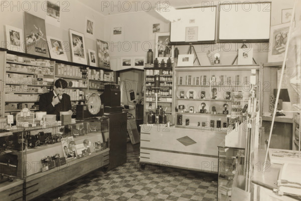 Interior of the Camera Shop in White Plains, New York, photograph, inscription: 'Camera Shop, probably 1938-39', United States of America, 1938 - 1939, photographer, printer (person), North America, paper, gelatin silver print, developing gelatin silver print, height 82 mm x width 128 mm, White Plains, A sepia-toned retail interior showing display counters, shelving, and wall-mounted posters. A man in a dark suit stands behind a glass-fronted counter filled with cameras and boxed items, holding something near his mouth and looking down at papers on the counter. Shelves behind him are stacked with small boxed goods and cylindrical containers, and framed pictures and promotional posters are arranged along the upper wall. A large round scale and a cash register sit on a counter near the man. Central shelving holds bottles and small jars, with additional display shelves showing various cameras and circular reflectors. A wooden sales counter with a dark diamond-shaped inlay faces the camera...