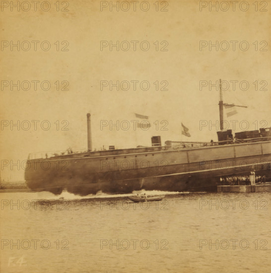 Launching of the HNLMS Hertog Hendrik at the Royal Shipyard in Amsterdam, The launching of a ship, Views and landscapes in Europe, Africa and Asia, Geographical Sculpture Gallery, collected by MJ Broekhuijsen, Head of a school in Amsterdam, stereograph, Numbered lower left of photo: F.4., annotation: 'The 'Hertog Hendrik'. See series A. No. 11 and series F. No. 3.', Amsterdam, publisher: Merkelbach & Co., compiler (person in literature): Martien Johan Broekhuijsen, 1902-06-07 - c. 1905, photographer, compiler (person in literature), pasteboard, paper, albumen print, height 87 mm x width 176 mm, Part of a group of 52 stereophotos used in primary education as a supplement to school charts. The entire series consisted of 274 items in 10 categories (A to K)., launching a ship, Rijkswerf (Amsterdam), A large ship hull fills the upper portion of the image with a smokestack and a mast rising above its deck, several flags hanging and waving from the mast, and a small boat floating on the water...