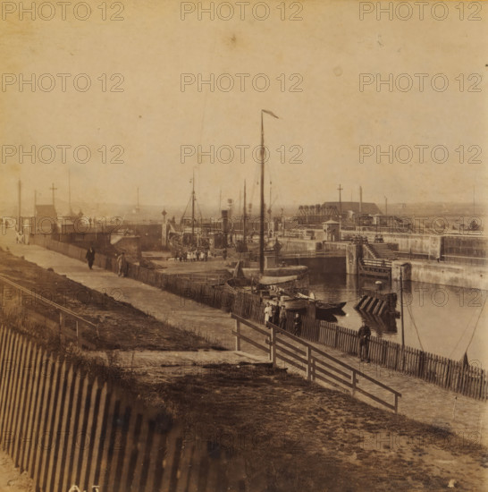 IJmuiden, View of IJmuiden southeast of the harbor, Views and landscapes in Europe, Africa and Asia, Geographical Sculpture Gallery, collected by MJ Broekhuijsen, Headmaster of a school in Amsterdam, stereograph, Numbered lower left of photo: A.5., annotation: 'We are standing southeast of the harbor. A boat is currently being locked through the lock.', publisher: Merkelbach & Co., Amsterdam, compiler (person in literature): Martien Johan Broekhuijsen, c. 1905, photographer, compiler (person in literature), pasteboard, paper, albumen print, height 86 mm x width 176 mm, Part of a group of 52 stereophotos used in primary education as a supplement to school charts. The entire series consisted of 274 numbers in 10 categories (A to K)., city-view in general, 'veduta', traffic on the water, A sepia-toned waterfront scene with a fenced pathway running alongside a harbor area where several small boats and larger vessels are moored; wooden piers, posts, and a tall mast with a small flag rise fr...