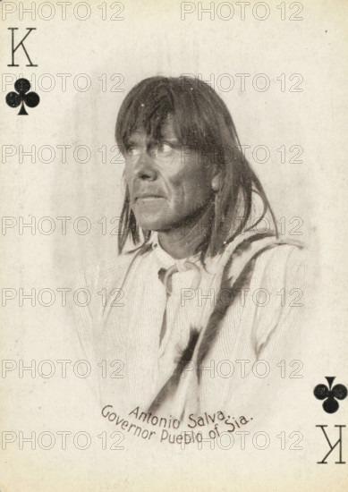 King of Clubs with portrait of Antonio Salva, Governor of Zia Pueblo, New Mexico, Antonio Salva. Governor Pueblo of Sia, photograph, playing card, Playing card from a box of 53 playing cards with autotype images of Native Americans. The contents include a complete set of playing cards including a joker., photographer: Adam Clark Vroman, United States of America, publisher: Lazarus & Melzer, Los Angeles (city), c. 1900, North America, paper, autotype, height 88 mm x width 63 mm, A sepia-toned portrait of a person shown from the chest up facing slightly to the viewer's left, with medium-length straight hair and a fringe partially covering the forehead, wearing a light-colored garment with a tied scarf at the neck and a darker strap or sash across one shoulder, the face marked by textured skin and a contemplative expression as the eyes look upward and to the left, set against a plain mottled background with dark club suit symbols in two corners.