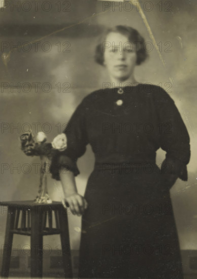 Rapid photographs, Snelfoto, photographer: Jan Gottfried Stütner, Alkmaar, 1913-07-09 - 1926, paper, gelatin silver print, developing gelatin silver print, height 74 mm x width 51 mm, A faded black-and-white portrait of a young woman standing with one hand resting on a small round-top stool; she wears a dark dress with puffed sleeves and a rounded neckline fastened by two circular brooches, and her hair is short and waved at the sides; a small vase with roses sits on the stool near her hand, and the background shows a softly mottled backdrop with visible scratches and creases on the photograph surface.