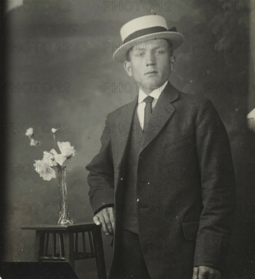 Rapid photographs, Snelfoto, photographer: Jan Gottfried Stütner, Alkmaar, 1913-07-09 - 1926, paper, gelatin silver print, developing gelatin silver print, height 66 mm x width 60 mm, A black and white portrait of a man wearing a three-piece suit and a boater hat with a dark band, standing beside a small wooden table on which a tall glass vase holds several light-colored flowers, the man’s hand resting on the table edge and his other hand relaxed at his side, with a plain, slightly mottled backdrop and some visible surface scratches on the photograph.