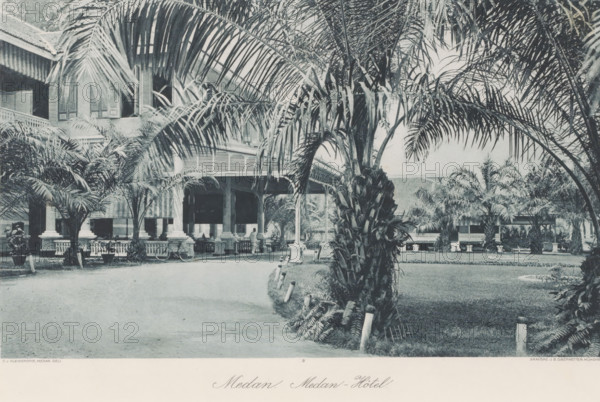 Medan. Medan Hotel, Garden and veranda of the Medan Hotel, photomechanical print, Part of Album with heliogravures of Sumatra's east coast., number: '9', photographer: Carl J. Kleingrothe, Medan, cliché maker: Johann Baptist Obernetter, Munich, in or before 1898, Carl J. Kleingrothe, Johann Baptist Obernetter, photographer, cliché maker, Bavaria, Sumatra, paper, photoaquatint, heliogravure, height 176 mm x width 290 mm, garden, trees: palm-tree, loggia, verandah, porch, A large building with a covered veranda is partly obscured by tall palm trees and other tropical foliage, with a wide driveway curving past manicured grass and planted borders, ornate columns and railings supporting the veranda, visible shuttered windows on the upper floor, a few potted plants and small decorative posts lining the lawn edge, and a person standing near the veranda under its shaded archways.