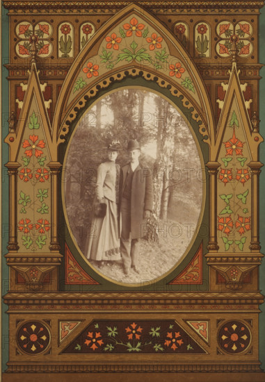 Portrait of a man and a woman in a forest, album page, photograph, cabinet photograph, This photo is part of an album., 1850 - 1900, photographer, paper, pasteboard, albumen print, height 136 mm x width 96 mm, historical persons, historical persons - BB - woman, forest, wood, A vertically oriented ornate frame with a pointed arch motif surrounds an oval photograph; the frame is decorated with stylized floral patterns in muted reds, greens, and gold tones, with symmetrical panels, columns, and carved-looking details; inside the oval, a sepia-toned portrait shows a man and a woman standing outdoors on grass in front of trees, the woman wearing a long skirt, fitted jacket, and a hat with a decorative element while holding a muff, and the man wearing a long overcoat, vest, trousers, and a bowler-style hat, both facing the camera and positioned close together.