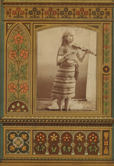 Portrait of a girl playing the violin, album page, photograph, cabinet photograph, This photo is part of an album., 1850 - 1900, photographer, paper, pasteboard, albumen print, height 136 mm x width 96 mm, historical persons - BB - woman, violin, fiddle, girl (child between toddler and youth), A sepia photograph of a young girl standing and playing a violin is centered within a decorative, floral border. She wears a striped dress with a sash tied at her waist and a patterned headscarf draped over her hair, and bracelets on her wrists. The girl looks slightly upward and holds the violin under her chin with one hand and the bow with the other. The background behind her is plain and light, with a curtain visible at one side and a fur rug at her feet. The surrounding frame features symmetrical ornamental panels with stylized flowers, leaves, geometric motifs, and warm earthy colors, including shades of brown, green, and orange, forming arch shapes and repeating floral patterns.
