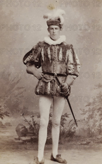Portrait of a young man, identified as AL van Haaften, album page, photograph, carte-de-visite, This photo is part of an album., inscription: 'Ant. J. van der Stok, PHOTOGRAPHE, Leiden Rapenburg 13.', name: 'AL van Haaften., 44.', photographer: Antonius Joannes van der Stok, Leiden, 1887-01-01 - 1899-12-03, paper, pasteboard, albumen print, albumendruk, height 83 mm x width 52 mm, historical persons, historical costume, A full-length sepia portrait of a young person standing in a studio setting wearing a historical costume with a puffed, patterned doublet and short padded trunk hose, a ruffled collar, and a small hat topped with a plume of feathers; one hand rests on the hip while the other holds a sword by its hilt, the sword pointing downward; the subject wears light-colored tights and ornate slippers; the backdrop shows painted foliage, rocks, and a slightly mottled background.