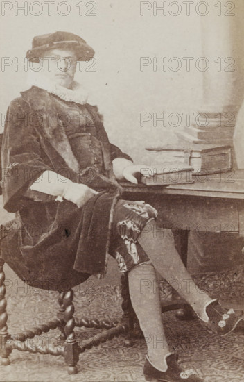 Portrait of a man in a suit sitting at a table, identified as N. Dirkzwager, album page, photograph, carte-de-visite, This photo is part of an album., name: 'N. Dirkzwager. 49', inscription: 'J. Goedeljee, Photographer, HRH PRINCE ALEXANDER, of the Netherlands, PRINCE of ORANGE, and HRH, PRINCESS HENDRIK, Hoogewoerd 160, near the Plantage.', photographer: Jan Goedeljee, Leiden, 1860 - 1905, paper, pasteboard, albumen print, height 83 mm x width 52 mm, historical persons, table, historical costume, A sepia portrait of a seated person wearing a dark velvet coat with fur trim and a ruffled white collar, a wide brimmed hat, patterned knee breeches, and decorative slippers, sitting on a carved wooden chair with twisted legs and resting one hand on a stack of books placed on a wooden table beside them.
