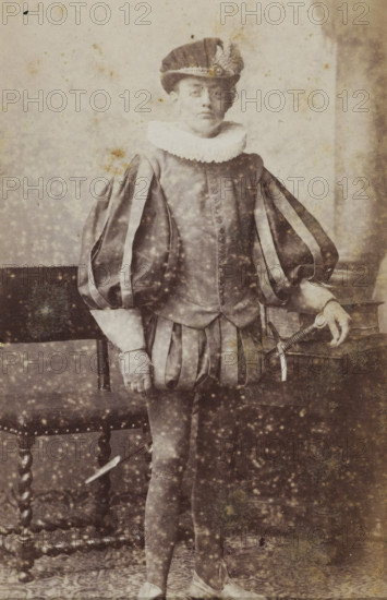 Portrait of a young man in costume, identified as WL Luijken Glashorst, album page, photograph, carte-de-visite, This photo is part of an album., name: 'WL Luijken Glashorst', inscription: 'J. Goedeljee, Photographer, HRH PRINCE ALEXANDER of the Netherlands, PRINCE of ORANGE, and HRH PRINCESS HENDRIK of the Netherlands, Hoogewoerd 160, near the Plantage.', photographer: Jan Goedeljee, Leiden, 1860 - 1905, paper, pasteboard, albumen print, height 83 mm x width 52 mm, historical persons, historical costume, A sepia-toned portrait of a person standing beside a wooden chair and a table, wearing a historical costume with a pleated ruff collar and a hat decorated with a feather, a fitted doublet with prominent puffed and striped sleeves, a skirted trunk hose, tights, and flat shoes; one hand rests on the table near a sword with a detailed hilt while the other hand hangs by the side, and the background shows a plain wall and a patterned floor with visible age spots and blemishes across the im...