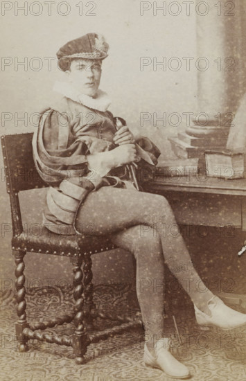 Portrait of a man in a suit sitting at a table with books, identified as J. Wiarda, album page, photograph, carte-de-visite, This photo is part of an album., inscription: 'J. Goedeljee, Photographer, HRH PRINCE ALEXANDER, of the Netherlands, PRINCE of ORANGE, and HRH, PRINCESS HENDRIK, Hoogewoerd 160, near the Plantage.', name: 'J. Wiarda, 52.', photographer: Jan Goedeljee, Leiden, 1860 - 1905, paper, pasteboard, albumen print, height 83 mm x width 52 mm, historical persons, historical costume, A seated young person in a historical costume with a brimmed hat adorned with trim and a ruffled collar looks toward the camera. They wear a doublet with slashed sleeves revealing inner fabric and fitted hose, and hold a slender object across their lap with both hands. The chair has a studded leather seat and twisted wooden legs, and a table beside them holds a closed book and an open book resting on its side. A patterned floor covering extends beneath the chair and their feet, and a column deta...