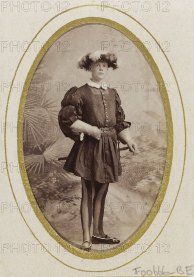 Portrait of a young man in costume with sword, referred to as CGJB Henny, album page, photograph, carte-de-visite, This photo is part of an album., name: 'CGJB Henny, 79.', inscription: 'Ant. J.van der Stok, PHOTOGRAPHE, Leiden Rapenburg 13.', photographer: Antonius Joannes van der Stok, Leiden, 1887-01-01 - 1899-12-03, paper, pasteboard, albumen print, albumendruk, height 83 mm x width 52 mm, historical persons, historical costume, hacking and thrusting weapons: rapier, An oval sepia photograph framed by a textured paper mat with a gilded inner border shows a person standing full length on a painted studio backdrop with foliage. The person wears a short dark tunic with a row of buttons down the front and a white collar, and the tunic has voluminous puffed sleeves with gathered sections. A wide belt or sash is tied at the waist and the skirt of the tunic falls above the knees. The person wears vertically striped stockings and pointed shoes with decorative elements on the toes. A wide-b...