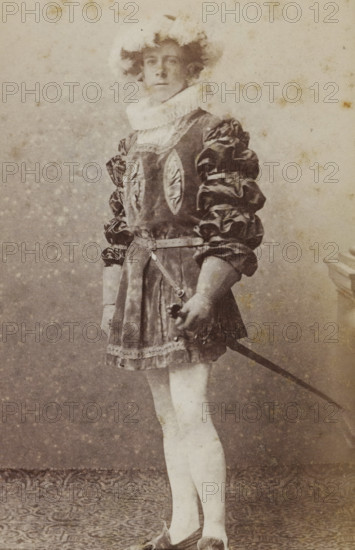 Portrait of Jan Gerritzen in costume with sword, album page, photograph, carte-de-visite, This photo is part of an album., name: 'J. Gerritzen., 102.', inscription: 'J. Goedeljee, Photographer, HRH PRINCE ALEXANDER, of the Netherlands, PRINCE of ORANGE, and HRH, PRINCESS HENDRIK, Hoogewoerd 160, near the Plantage.', photographer: Jan Goedeljee, Leiden, 1886 - 1892, paper, pasteboard, albumen print, albumendruk, height 83 mm x width 52 mm, Person, Jan Gerritzen, historical persons, historical costume, hacking and thrusting weapons: rapier, A faded sepia photograph of a person standing in a full-length pose wearing a costume with a short skirt and puffed sleeves, a ruffled high collar around the neck, and a belt with a strap crossing the waist; the left hand holds the hilt of a sword whose blade extends downward and out of frame, while the right hand rests near the hip; the person has light-colored stockings and pointed shoes, and a hat or headpiece with fluffy details sits atop tousled ...