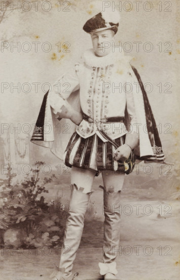 Portrait of a young man in costume with sword, referred to as A. Ypey, album page, photograph, carte-de-visite, This photo is part of an album., name: 'A. Ypey., 183.', inscription: 'Ant. J. van der Stok, PHOTOGRAPHE, Leiden Rapenburg 13.', photographer: Antonius Joannes van der Stok, Leiden, 1887-01-01 - 1899-12-03, paper, pasteboard, albumen print, albumendruk, height 83 mm x width 52 mm, historical persons, historical costume, hacking and thrusting weapons: rapier, A full-length portrait of a person standing in a patterned studio setting wearing an ornate historical costume with a ruffled collar, a decorated sleeved jacket, a short pleated skirt with vertical stripes, and a cape draped over the shoulders; the person wears a hat with a plume, holds a mask or small object in one hand, and has one hand resting on their hip; visible legs are covered in fitted stockings with decorative ties and shoes featuring bows; the background shows painted foliage and a faint decorative gate, and th...