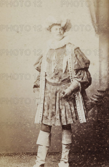 Portrait of a young man in costume, identified as BPG van Diggelen, album page, photograph, carte-de-visite, This photo is part of an album., inscription: 'J. Goedeljee, Photographer, HRH PRINCE ALEXANDER of the Netherlands, PRINCE of ORANGE, and HRH PRINCESS HENDRIK of the Netherlands, Hoogewoerd 160, near the Plantage.', name: 'BPG v. Diggelen., 247.', photographer: Jan Goedeljee, Leiden, 1860 - 1905, paper, pasteboard, albumen print, height 83 mm x width 52 mm, historical persons, historical costume, A sepia-toned photograph of a person standing and facing slightly to the side, wearing a historical costume with a ruffled collar and a decorative bodice, puffed sleeves and long ornate ribbons hanging from the shoulders down the front of the outfit, a pleated skirt with lace trim and a patterned apron-like overlay, tall layered boots covering the calves, and a hat with a feather.
