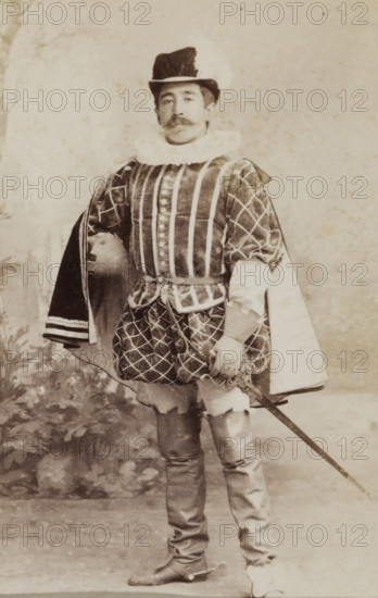 Portrait of a young man in costume with sword, identified as W. Birnie, album page, photograph, carte-de-visite, This photo is part of an album., inscription: 'Ant. J. van der Stok, PHOTOGRAPHE, Leiden Rapenburg 13.', name: 'W. Birnie 249.', photographer: Antonius Joannes van der Stok, Leiden, 1887-01-01 - 1899-12-03, paper, pasteboard, albumen print, albumendruk, height 83 mm x width 52 mm, historical persons, hacking and thrusting weapons: sword, historical costume, A sepia-toned photograph of a man standing and facing forward wearing a ruffled collar and a tall hat with a brim. He has a mustache and is dressed in a patterned doublet with vertical stripes on the front and diamond-patterned sleeves, and a short, quilted skirt over fitted hose. One arm rests on his hip beneath a cloak draped over his shoulder, and the other hand holds the hilt of a sword that extends downward. He wears tall boots with turned-down tops and the background shows a plain, lightly mottled backdrop with fain...