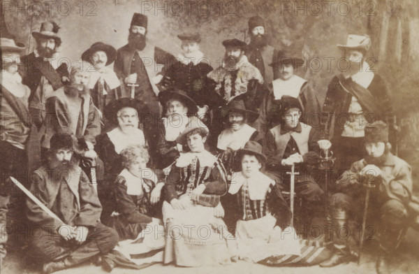 Group portrait of students in costume, album page, photograph, carte-de-visite, This photo is part of an album., inscription: 'J. Goedeljee, Photographer, HRH PRINCE ALEXANDER of the Netherlands, PRINCE of ORANGE, and HRH PRINCESS HENDRIK of the Netherlands, Hoogewoerd 160, near the Plantage.', photographer: Jan Goedeljee, Leiden, 1880 - 1900, paper, pasteboard, albumen print, height 83 mm x width 52 mm, student, anonymous historical persons portrayed in a group, in a group-portrait, historical costume, A sepia photograph showing a large group of people arranged in rows, many wearing period costumes with wide collars, hats, cloaks, and beards; some individuals hold swords and one person rests a hand on a cane; central figures include seated women in striped bodices and full skirts, surrounded by men in varied headwear from wide-brimmed hats to fur caps and tall hats, and a few figures wear fur-trimmed garments; expressions are mostly neutral, and the background is plain and softly text...