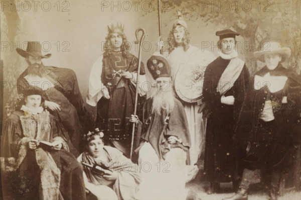 Group portrait of students in costume, album page, photograph, carte-de-visite, This photo is part of an album., inscription: 'J. Goedeljee, Photographer, HRH PRINCE ALEXANDER of the Netherlands, PRINCE of ORANGE, and HRH PRINCESS HENDRIK of the Netherlands, Hoogewoerd 160, near the Plantage.', photographer: Jan Goedeljee, Leiden, 1880 - 1900, paper, pasteboard, albumen print, height 83 mm x width 52 mm, student, anonymous historical persons portrayed in a group, in a group-portrait, historical costume, A sepia-toned group portrait showing several people in period or theatrical costumes arranged in two rows, with some seated and others standing; visible are ornate hats and headpieces including a crown, a tall mitre-like hat, and wide-brimmed hats, along with long robes, capes, fur-trimmed garments, and a variety of collars and sashes; one seated figure holds a staff and has a long beard, another standing figure holds a tall pole, a seated person holds an open book, and a reclining pers...