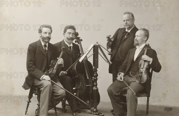 Group portrait of a string quartet, album page, photograph, cabinet photograph, This photo is part of an album., photographer: Wilhelm Höffert, 1901, paper, pasteboard, gelatin silver print, developing gelatin silver print, height 134 mm x width 196 mm, historical persons, adult man, small group of musicians, chamber orchestra, jazz band, A black-and-white photograph of four seated men in formal suits arranged around a central music stand, each holding a string instrument; two men hold violins with bows visible, one man holds a cello with its endpin on the floor, and one man holds a viola while resting a bow on his knee. The men have varied facial hair styles including mustaches and beards, and they wear dark jackets, waistcoats, and ties or cravats. The music stand has an open score visible on its ledge and slender metal supports. The chairs are wooden with turned legs and carved details. The background is plain and light-colored with some mottled spots and age marks on the image surf...