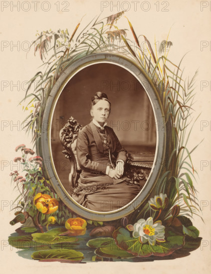 Portrait of a woman sitting on a chair at a table on which an album lies, photograph, cabinet photograph, This photo is part of an album., inscription: 'Photographischer Salon, von, Emil Tiedemann, 12. Richtweg 12., BREMEN, Für restellungen bleibt, die Platte aufbewahrt.', photographer: Emil Tiedemann, Bremen (city), 1859 - 1892, paper, pasteboard, albumen print, albumendruk, height 135 mm x width 96 mm, historical persons - BB - woman, chair, table, book, An oval sepia photograph of a seated woman framed by a painted floral and reed border, showing the woman in a dark high-collared dress with layered bodice and decorative buttons, a brooch at her throat, her hair styled in an updo, hands folded and resting on a fringed shawl over her lap and on an ornately carved wooden chair beside a table, while the surrounding painted frame includes tall grasses and reeds at the top and sides and colorful flowers and broad lily pads with a blooming water lily at the bottom.