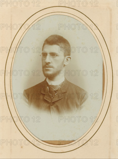 Portrait of a man with glasses, album page, photograph, cabinet photograph, This photo is part of an album., inscription: 'HW Wollrabe, COURT PHOTOGRAAF, The Hague, Noordeinde 96.', photographer: Heinrich Wilhelm Wollrabe, 1877 - 1901, paper, pasteboard, daylight collodion silver print, height 137 mm x width 97 mm, historical persons, eyeglasses, spectacles, A faded oval portrait of a man with short hair and a trimmed beard wearing round wire-rimmed glasses and a high-collared shirt with a patterned necktie and a pinstriped jacket, set against a muted pale background and bordered by concentric oval frames on an aged paper mount.