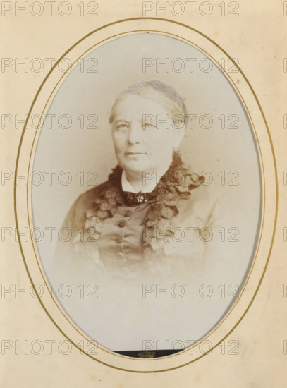 Portrait of a woman, album page, photograph, cabinet photograph, This photo is part of an album., inscription: ', Dupont, 67, Rue Neuve, Bruxelles, ', photographer: Dupont (photo studio), 1864 - 1886, paper, pasteboard, albumen print, height 134 mm x width 96 mm, historical persons, adult woman, An oval-framed sepia portrait of an older woman shown from the chest up, wearing a high-collared dress with a dark ruffled trim and a small decorative brooch at the throat, her hair pulled back and covered with a thin headband, her expression neutral, set against a pale, slightly mottled background and surrounded by a cream mat with concentric gold lines.
