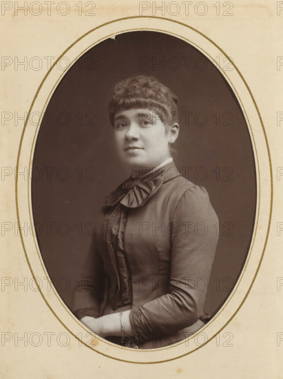 Portrait of a young woman, album page, photograph, cabinet photograph, This photo is part of an album., inscription: 'Inalterable Process, Charcoal Photography, Nestor Schaffers, Photographer, Rue Guinard 9, Ghent', inscription: 'Inalterable charcoal impression, photographer: Paul Joseph Nicolas Nestor Schaffers, 1875 - 1896, Paul Joseph Nicolas Nestor Schaffers, photographer, paper, pasteboard, carbon print, height 134 mm x width 96 mm, historical persons, adolescent, young woman, maiden, A sepia portrait of a young woman shown from the waist up inside an oval frame, wearing a high-collared dress with a large bow at the neck and decorative vertical ruffles down the front, her hair styled with short bangs and a curled updo, she faces slightly toward the camera with a neutral expression, her hands folded in her lap with a delicate bracelet visible on one wrist, the background inside the oval is plain and darker than the surrounding light-colored mat.