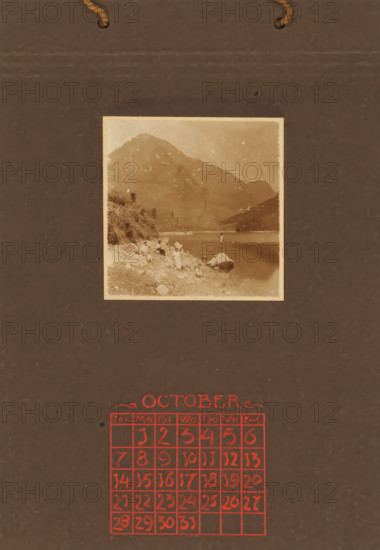 Three women, a man and two children on rocks on the coast, with a mountain in the background, album page, photograph, This photo is part of an album., annotation: 'OCTOBER, ', Indonesia, 1880 - in or before 1923, photographer, pasteboard, paper, daylight collodion silver print, height 80 mm x width 78 mm, adult man, mountains, rocky coast, child (two persons), adult woman (three persons), A sepia-toned photograph mounted on a dark brown textured background shows a lakeshore scene with mountains rising behind the water; the shoreline is rocky with grass and scattered stones, and several small figures in period clothing are positioned along the bank and on a large rock at the water's edge. The background surface has two short pieces of rope threaded through holes near the top and faint scuff marks and spots across the brown area. Below the photograph a grid of hand-drawn squares filled with hand-drawn numerals is printed in a contrasting color.