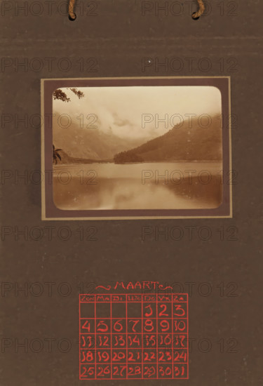 Landscape with mountains and lake, album page, photograph, This photo is part of an album., annotation: 'MARCH, ', Indonesia, 1880 - in or before 1923, photographer, pasteboard, paper, daylight collodion silver print, height 80 mm x width 110 mm, lake, mountains, A sepia photographic print of a misty lakeside scene mounted on a dark brown background with rounded inner corners, showing calm water in the foreground reflecting the sky, low-lying mist or clouds hugging distant mountains and hills, a small wooded peninsula jutting into the lake, and a few overhanging branches visible at the frame edge, with a rope loop visible near the top and a grid of hand-drawn squares rendered in bright red ink toward the lower portion of the background.