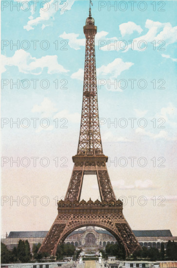 Paris. - La Tour Eiffel., View of the Eiffel Tower in Paris, photograph, page, Part of Photo album with color shots of sights in Paris., Paris, c. 1900 - c. 1930, photographer, pasteboard, paper, autotypie, color autotypie, height 197 mm x width 159 mm, traveling, tourism, prospect of city, town panorama, silhouette of city, Eiffel tower, A tall iron lattice tower rises from the bottom of the image into a sky filled with pale blue and soft white clouds, the structure narrowing as it reaches a domed top with a small flagpole. The tower shows ornate cross-braced metalwork and two broad horizontal platforms with decorative railings, and its base arches over a landscaped area with trees and formal pathways. Behind and beneath the arch there is a large classical-style building with an arched central facade and flanking wings, and rows of trees and garden elements lead toward the foreground. The overall palette is muted with reddish-brown tones for the tower, soft blues and whites for the sk...