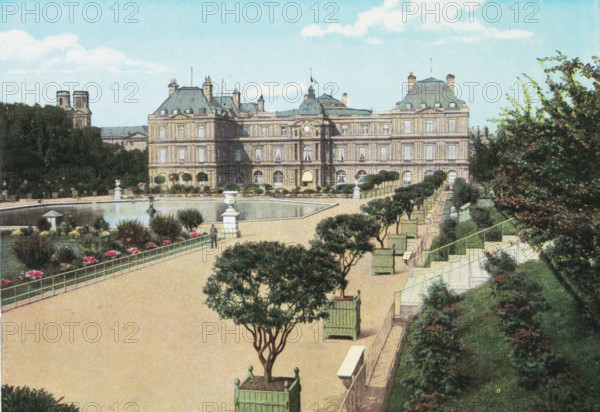 View of the Jardin du Luxembourg in Paris, Paris. - Le Jardin du Luxembourg., photograph, page, Part of Photo album with color shots of sights in Paris., Paris, c. 1900 - c. 1930, photographer, pasteboard, paper, autotypie, color autotypie, height 197 mm x width 159 mm, traveling, tourism, prospect of city, town panorama, silhouette of city, public gardens, park (city(-scape) with figures, staffage), Jardin du Luxembourg, A wide view of a formal garden with a long rectangular pond surrounded by a paved walkway, ornamental urns on pedestals, neatly trimmed small trees in square planters along the promenade, rows of green wooden chairs facing the central area, flowerbeds with bright blooms near the water, a person standing near the pond edge, dense trees and hedges framing the scene, and a large classical stone building with multiple windows, mansard roofs, chimneys, and a central clocked pavilion under a blue sky with scattered clouds.