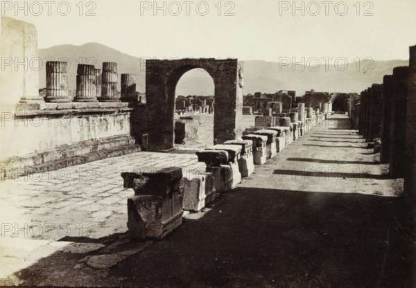 Foro e Tempio di Giove di Pompei., Remains of the forum and the Temple of Jupiter in Pompeii, album page, photograph, Part of Photo album with recordings of excavations in Pompeii by Roberto Rive., number: '407', photographer: attributed to Roberto Rive, Pompeii, c. 1860 - c. 1900, Attributed to, paper, albumen print, height 104 mm x width 141 mm, square, circus, temple, shrine, Roman religion, excavation, archaeology, A wide, sunlit scene of ancient stone ruins with a paved foreground and a long shadowed pathway running alongside a row of low rectangular stone blocks, a prominent arched stone gateway standing near the center, fluted column fragments on a raised platform to the left, scattered carved stone pieces leading toward distant ruined walls and structures, and low hills visible on the horizon under a pale sky.